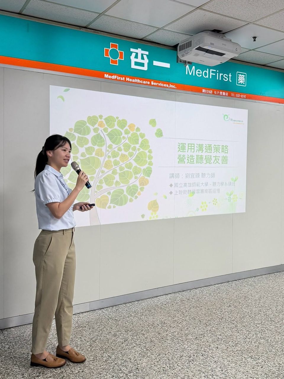 上聆聽力師在台大醫院候診區以麥克風進行衛教宣導，身後投影幕是「運用溝通策略 營造聽覺友善」主視覺